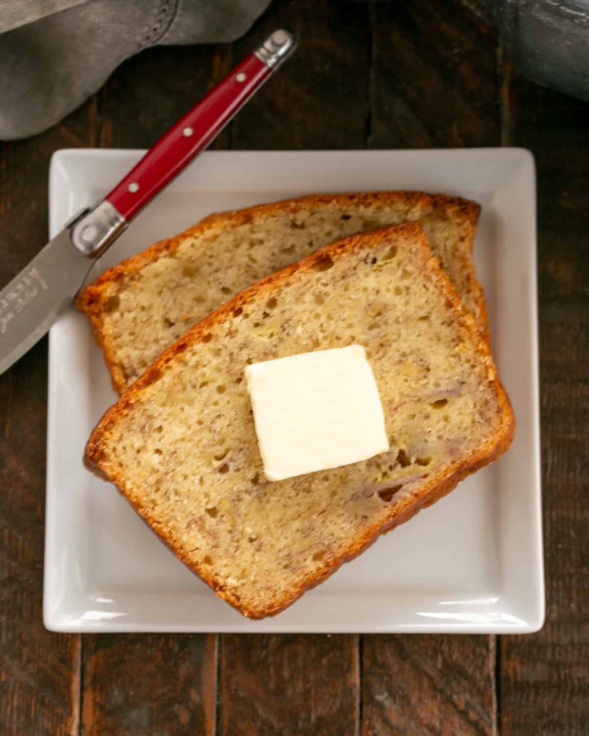 2 slices of banana bread with a pat of butter on the top on a square white plate with a red handled knife.