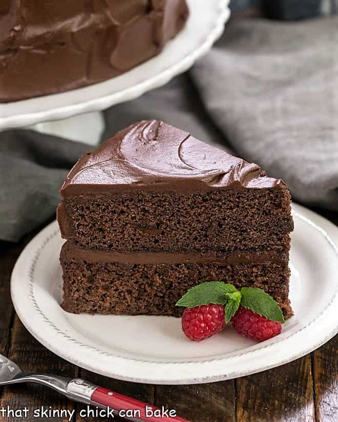 Slice of  Devil's Food Cake on a white dessert plate with a red handled fork with cake stand in the background.
