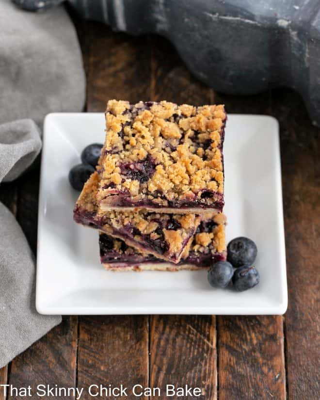 A stack of 3 blueberry bars on a square white dessert plate with fresh blueberries to garnish
