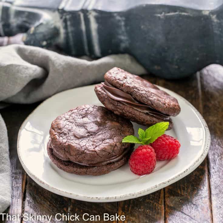 2 sandwich cookies on a round white plate with raspberry and mint garnish