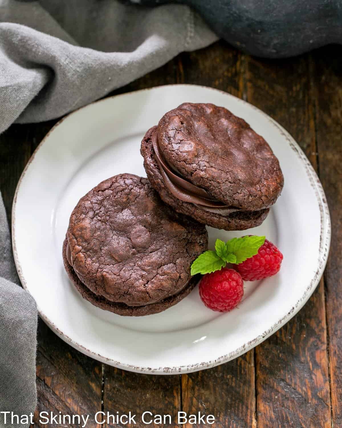 Overhead view of two chocolate sandwich cookies on a white dessert plate with 2 raspberries and a sprig of mint.